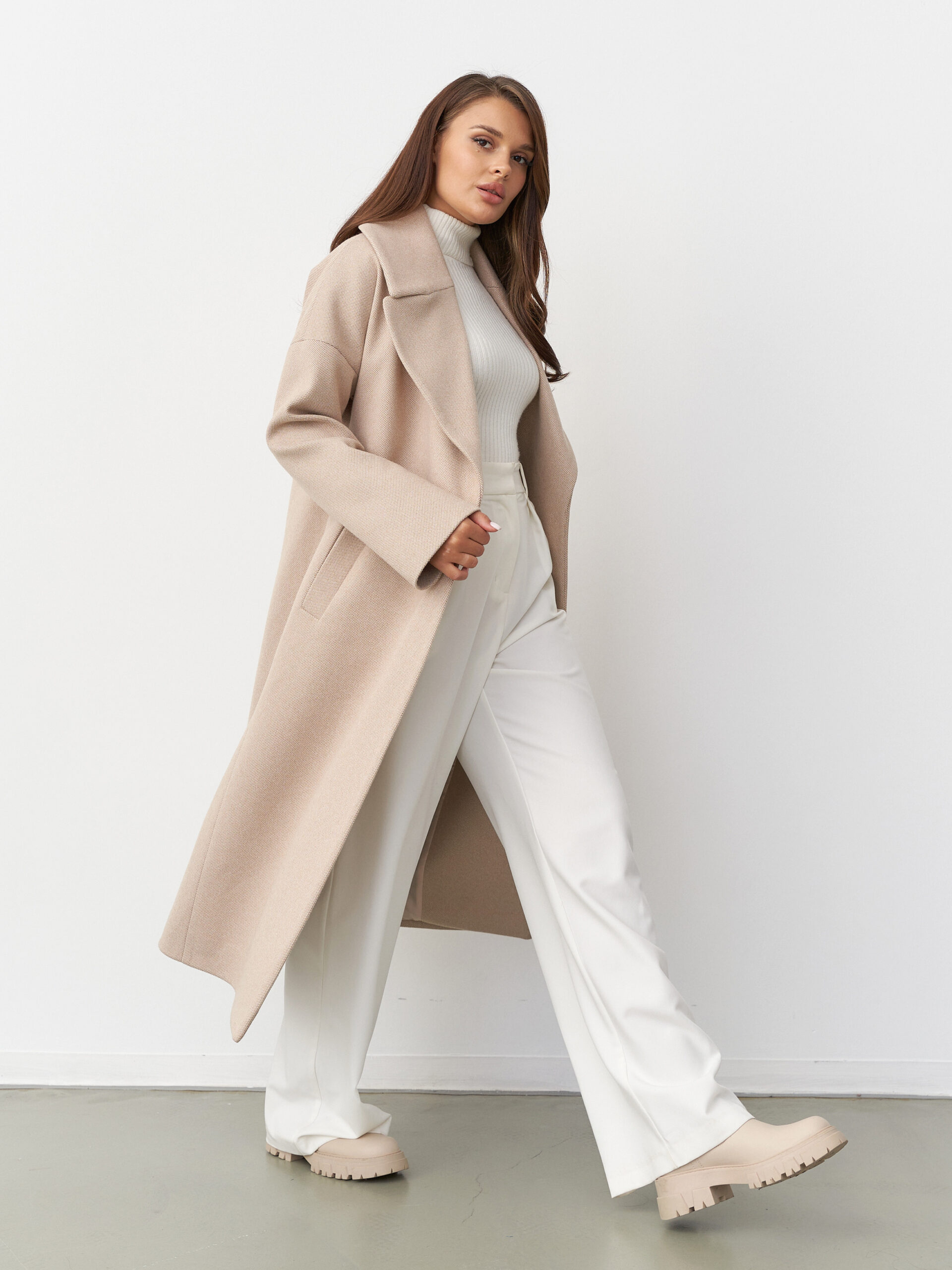 Stylish woman in beige coat, white turtleneck, and trousers walks confidently. Light brown hair, casual yet sophisticated look. Neutral tones enhance the elegant and modern outfit